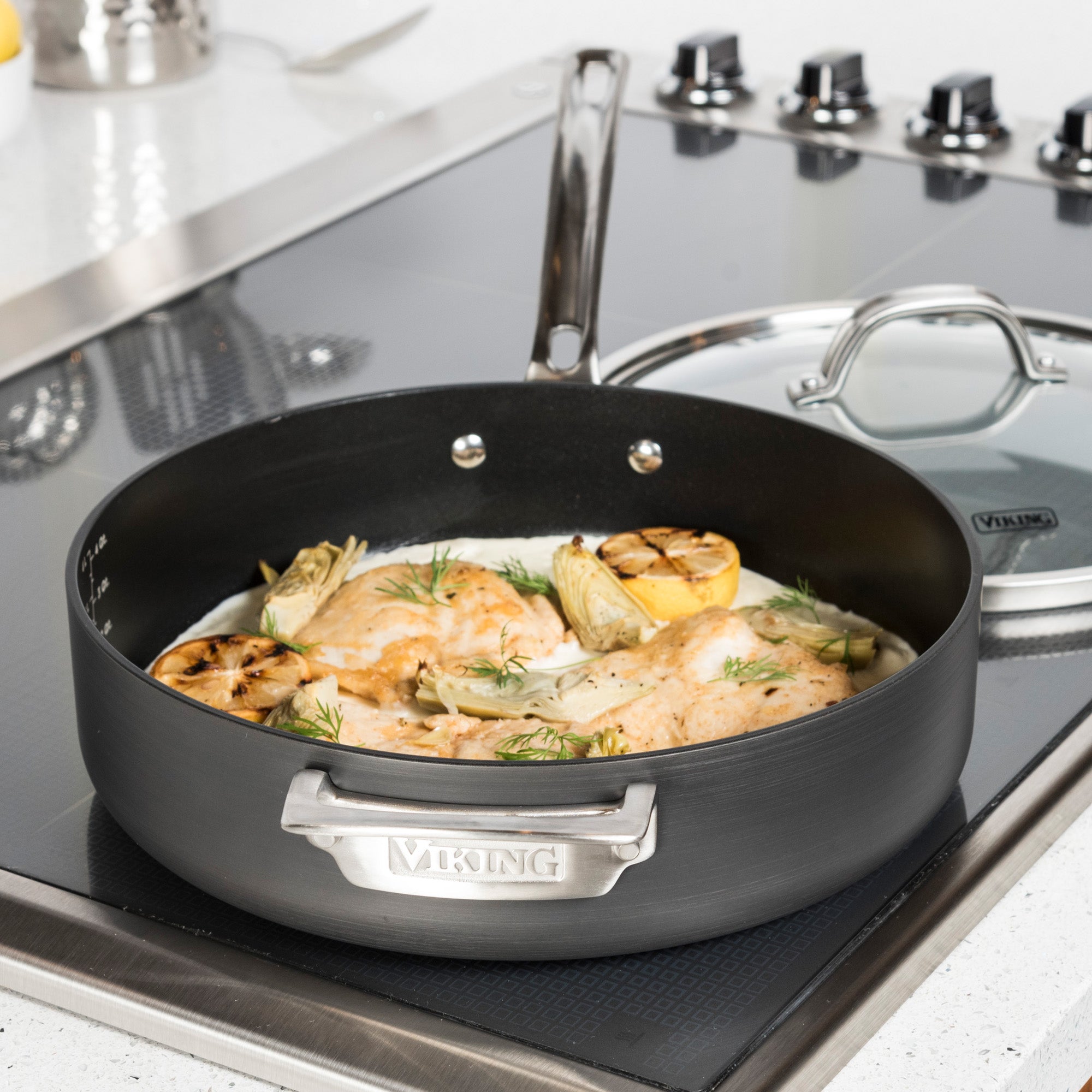 A Viking Culinary Products Hard Anodized Nonstick pan from the 10-Piece Cookware Set with Glass Lids sits on a stovetop, holding cooked chicken breasts, grilled lemons, herbs, and artichokes. A glass lid rests nearby in a modern kitchen.