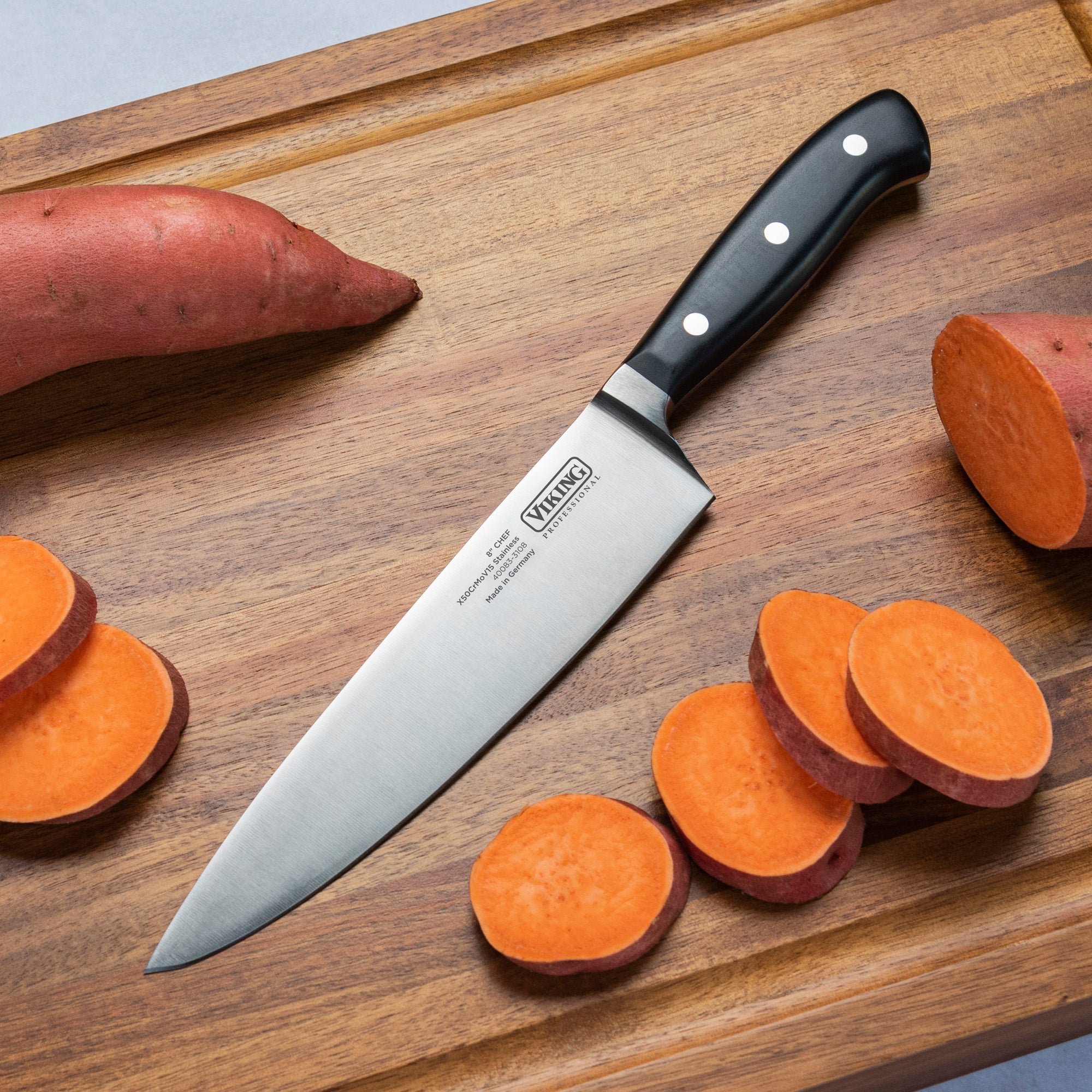 The Viking Professional 8-Inch Chefs Knife by Viking Culinary Products, featuring a full-tang design and black handle, rests on a wooden cutting board with whole and sliced sweet potatoes nearby.