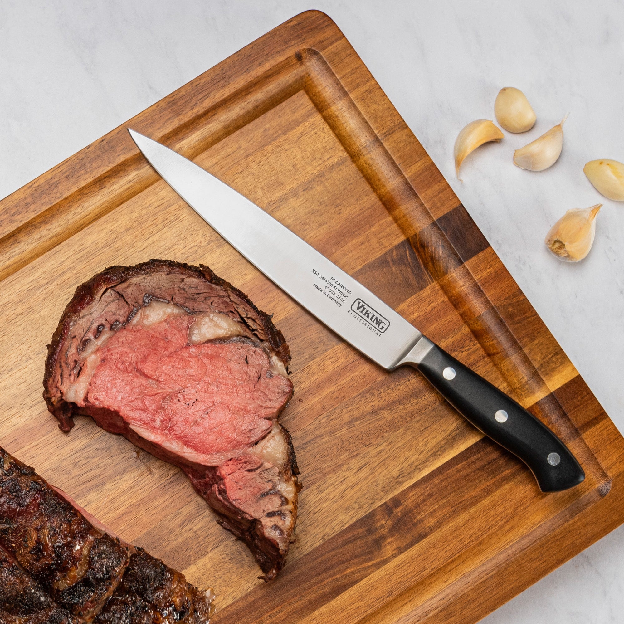 A Viking Professional 8 Carving Knife by Viking Culinary Products rests on a wooden cutting board with partially sliced roast beef and three garlic cloves, all on a white surface.