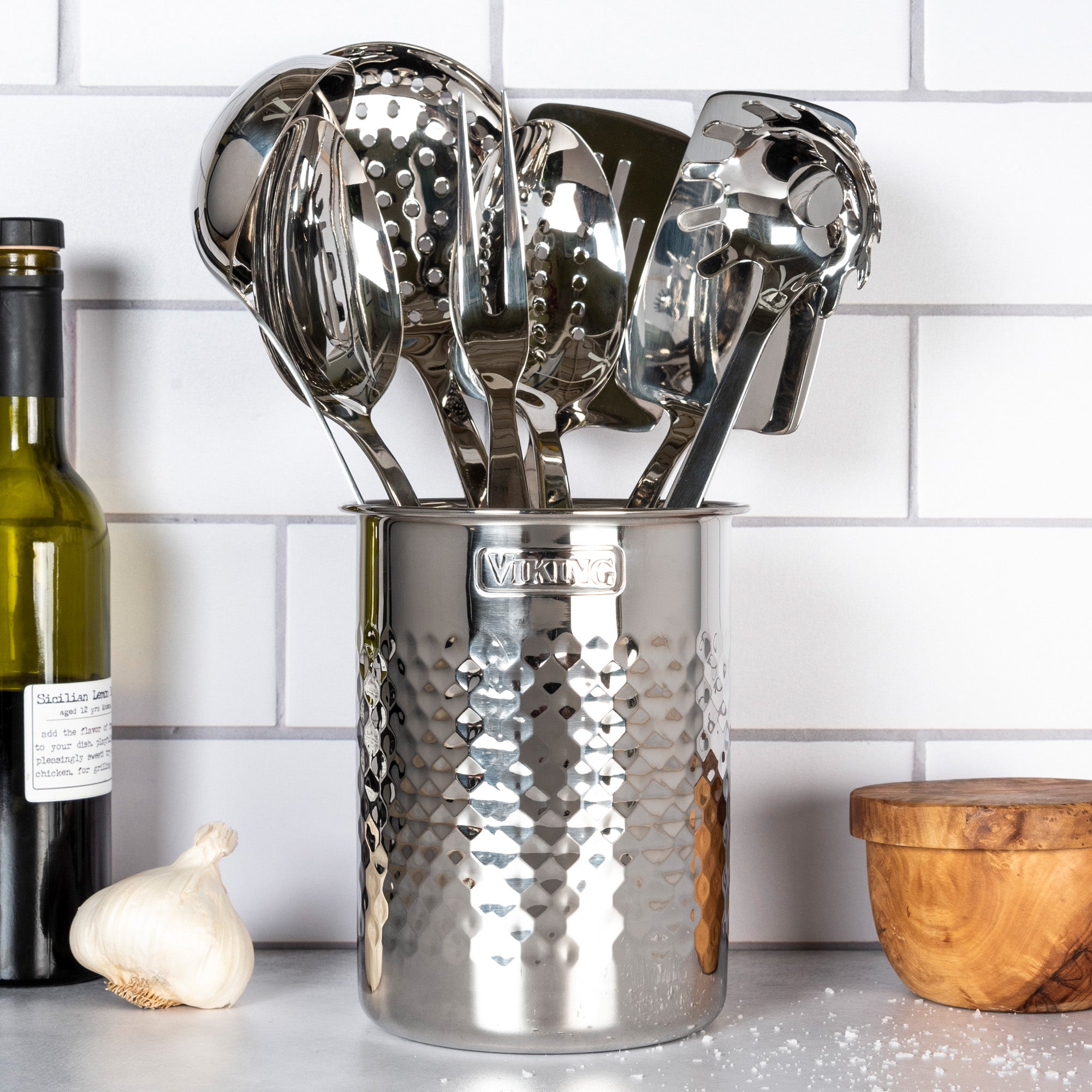 A set of shiny kitchen utensils is organized in the Viking Hammered Stainless Steel Utensil Holder by Viking Culinary Products, sitting beside olive oil, garlic, and a wooden container against a white tiled backsplash.