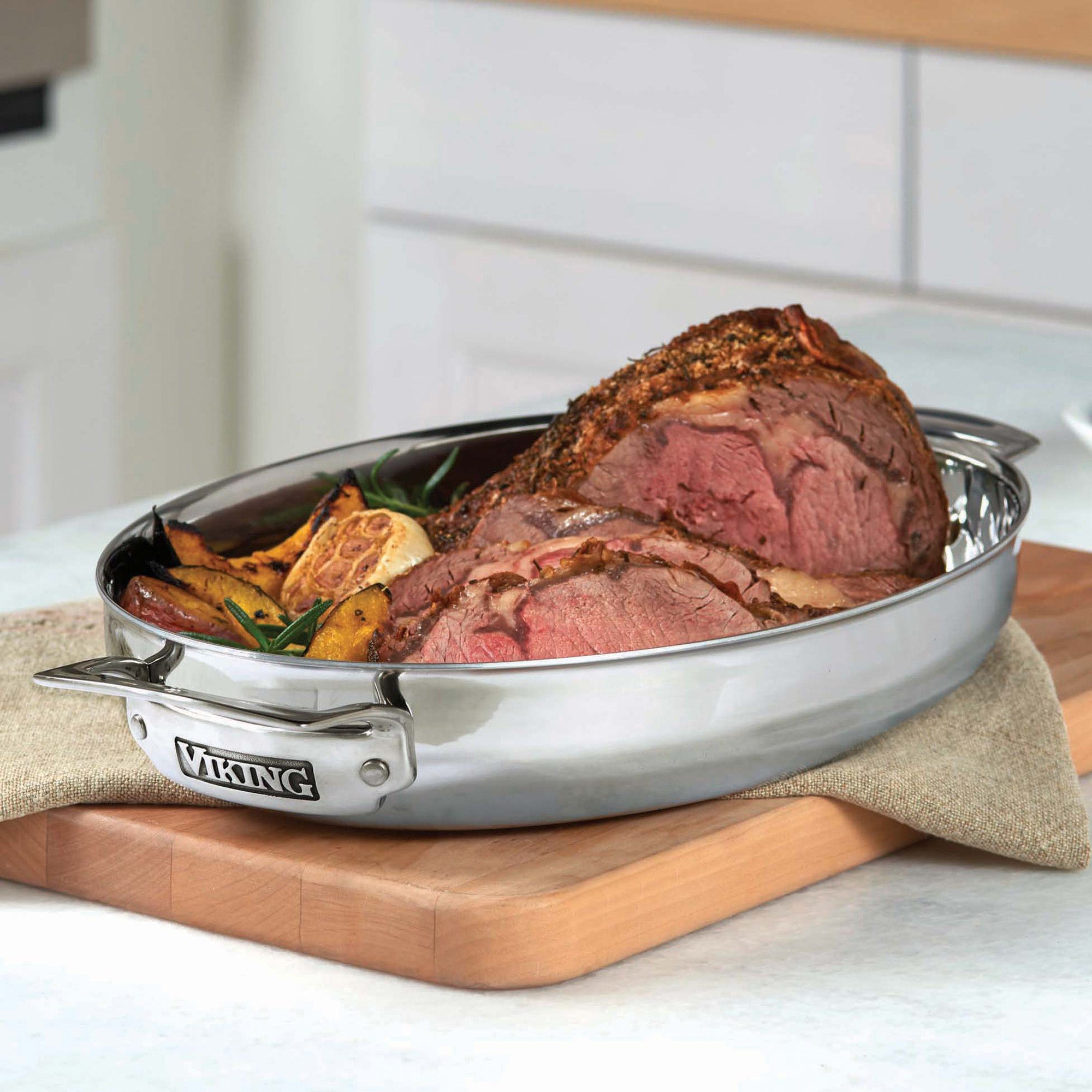 The Viking 3-Ply 8.5-Quart 3-in-1 Oval Roaster with Rack by Viking Culinary Products holds a sliced roast with herbs and potatoes, resting on a beige cloth in a modern kitchen. This induction-compatible pan offers versatile cooking.