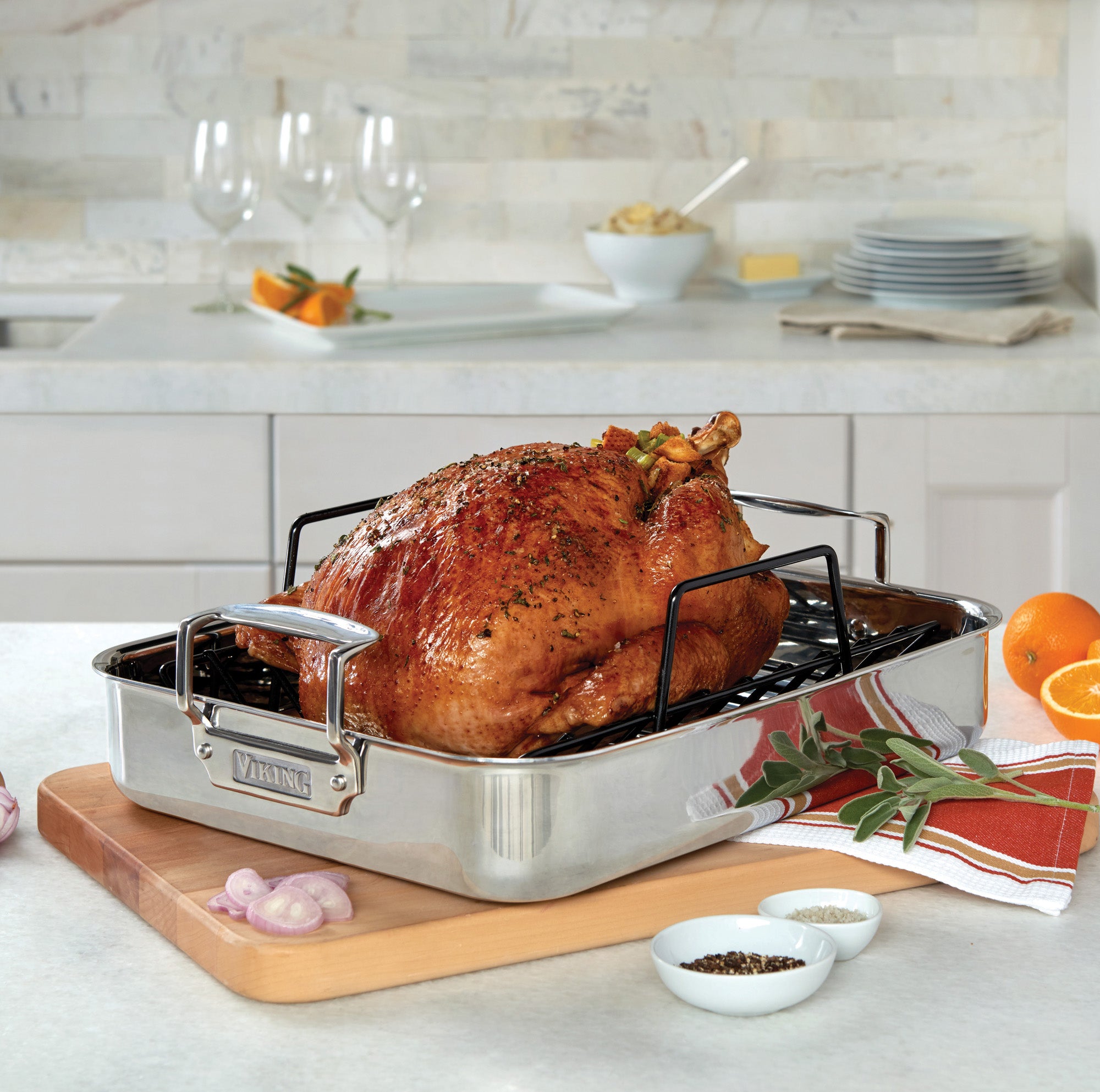A roasted turkey rests in the Viking 3-Ply Stainless Steel Roaster with Rack by Viking Culinary Products, garnished with herbs on a kitchen counter. Sliced oranges, sage, shallots, salt, pepper, wine glasses, and a bowl are nearby.