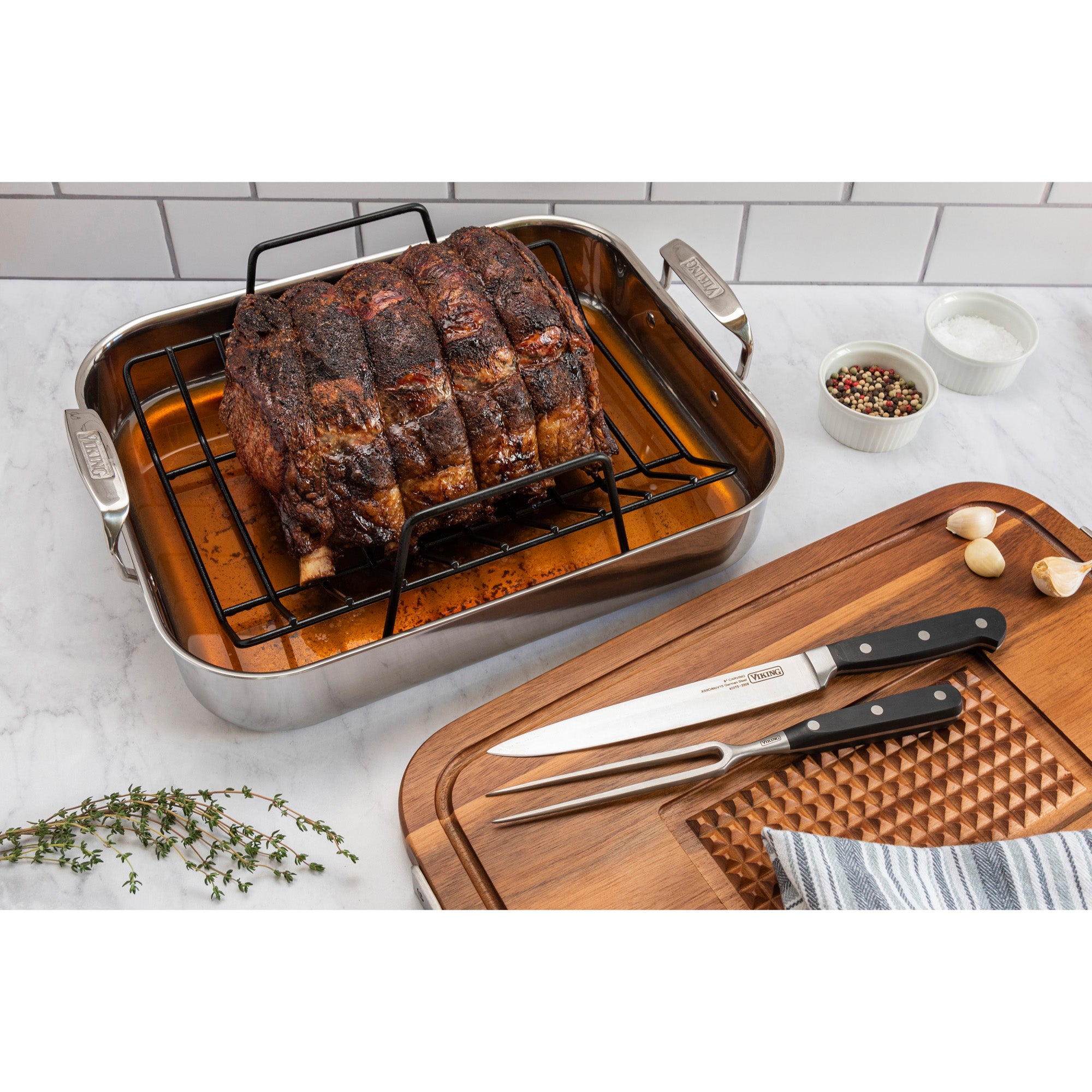 A beef rib roast rests on the Viking 3-Ply Stainless Steel Roaster with Rack and Bonus Carving Set by Viking Culinary Products, accompanied by thyme, garlic, a striped towel, peppercorns, salt bowls, and a cutting board.