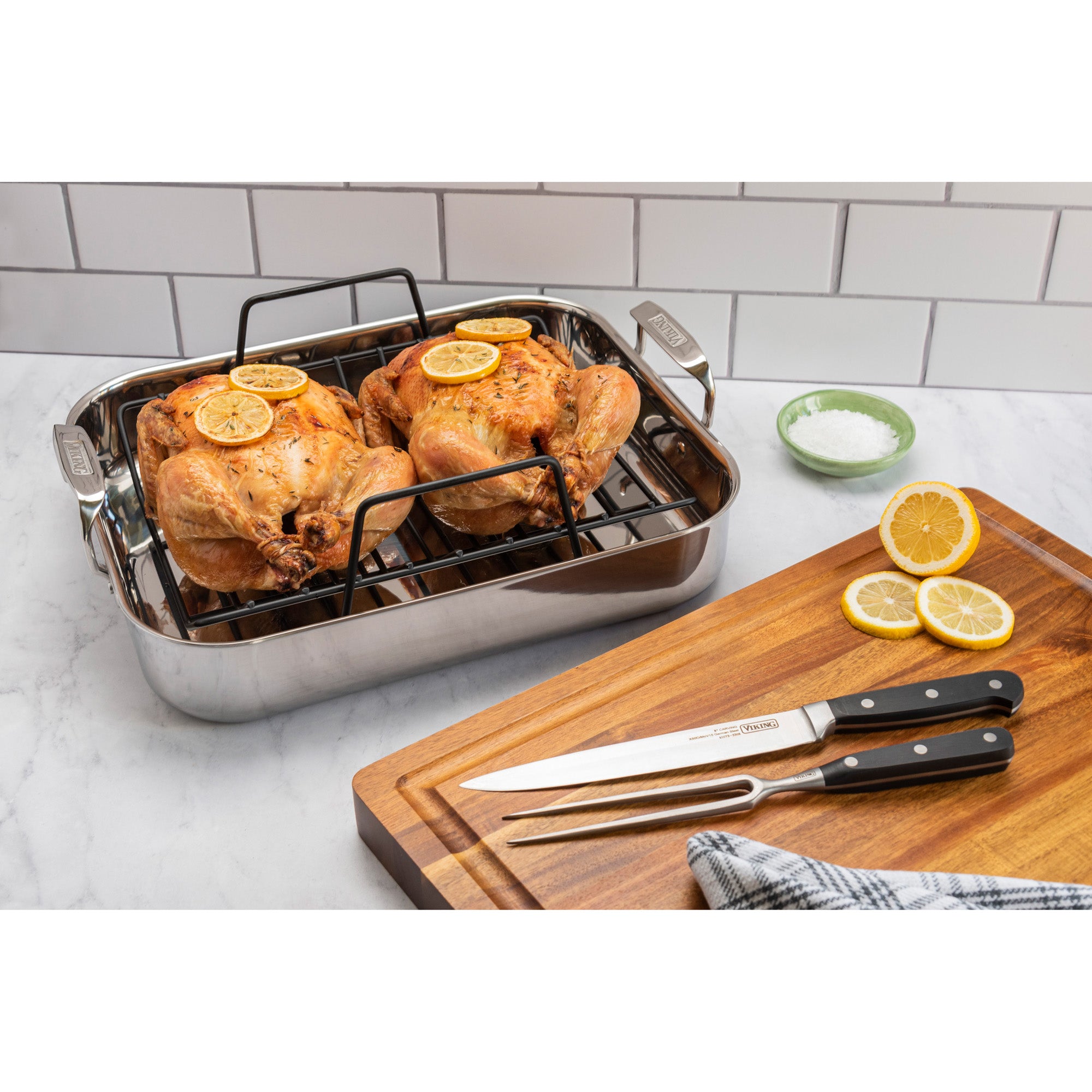 Two roasted chickens with lemon slices rest in a Viking 3-Ply Stainless Steel Roaster with Rack by Viking Culinary Products. A wooden board holds the bonus carving set, extra lemon slices, and a bowl of salt nearby.