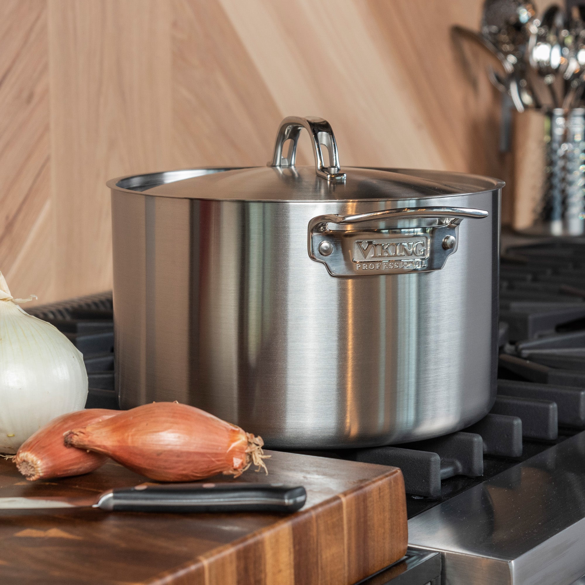 A Viking Professional 5-Ply Stainless Steel 8-Quart Stock Pot with lid from Viking Culinary Products sits on the stove. In front, a cutting board holds a knife, an onion, and two shallots; kitchen utensils appear in the background.
