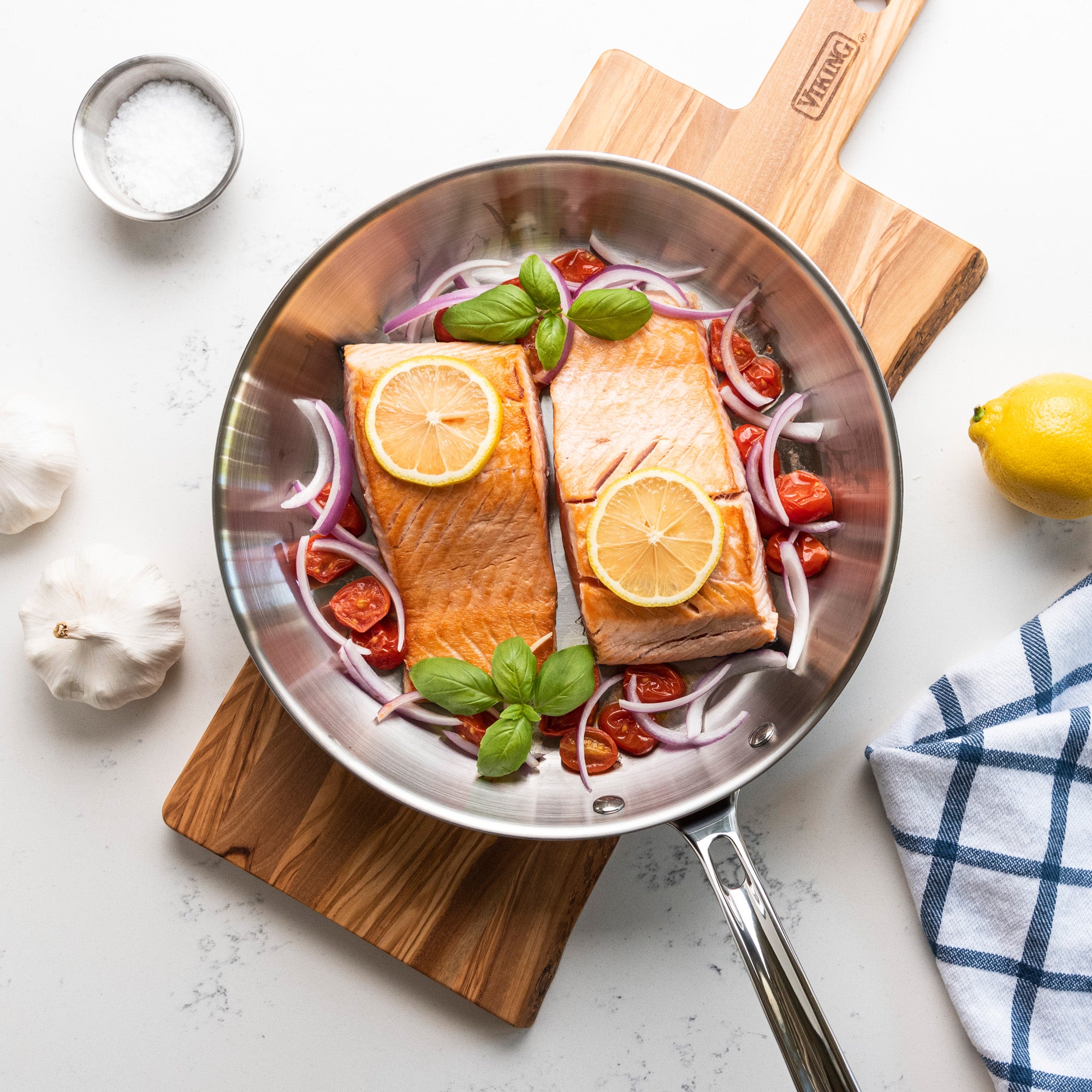 The Viking Professional 5-Ply 10-Inch Fry Pan from Viking Culinary Products holds two salmon fillets with lemon slices, cherry tomatoes, red onion, and basil. Nearby: garlic bulbs, a lemon, salt, and a kitchen towel.