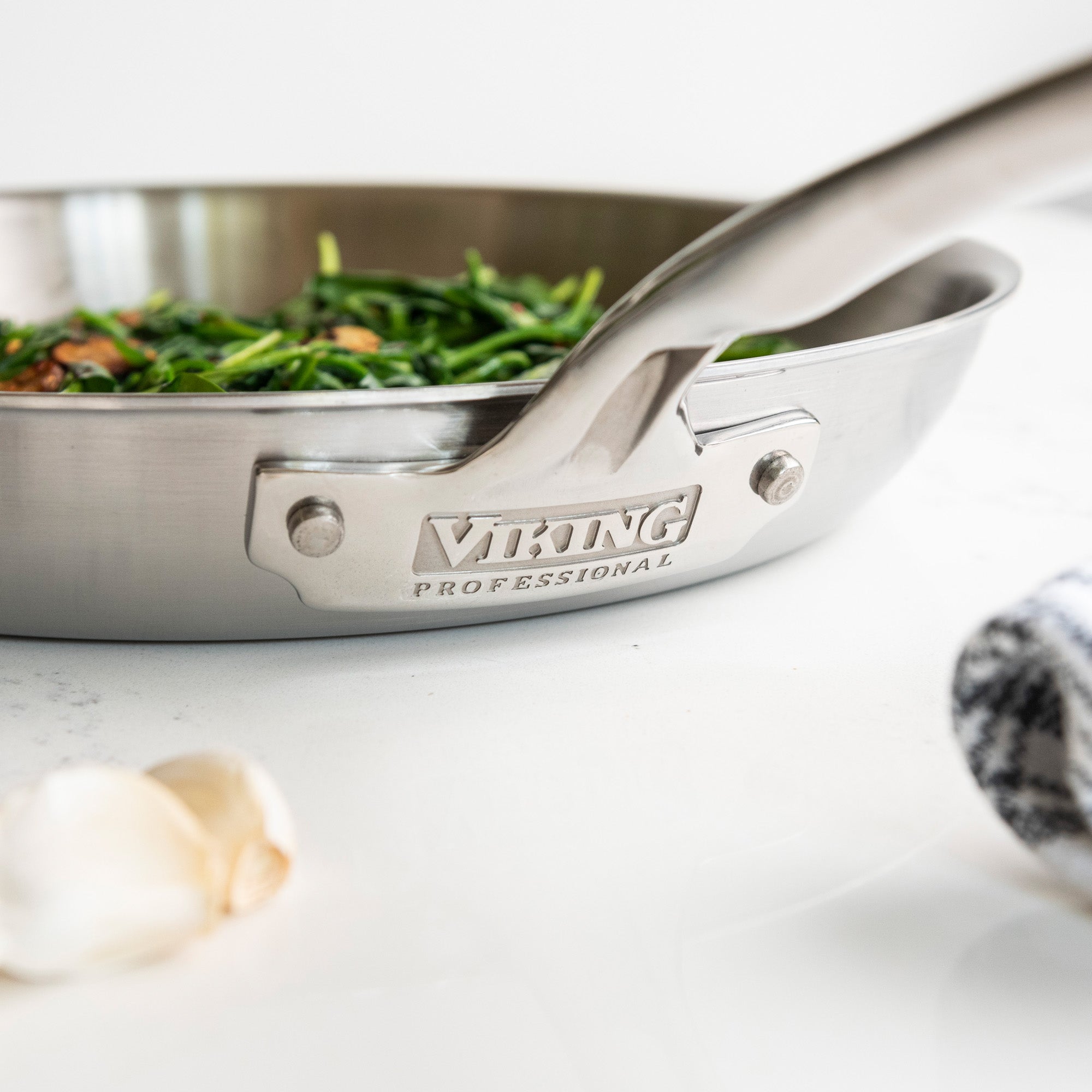 A Viking Culinary Products Professional 5-Ply 12-Inch Fry Pan, filled with sautÃ©ed greens, sits on a white surface near garlic cloves and a striped towel. This skillet is dishwasher safe for easy cleanup.