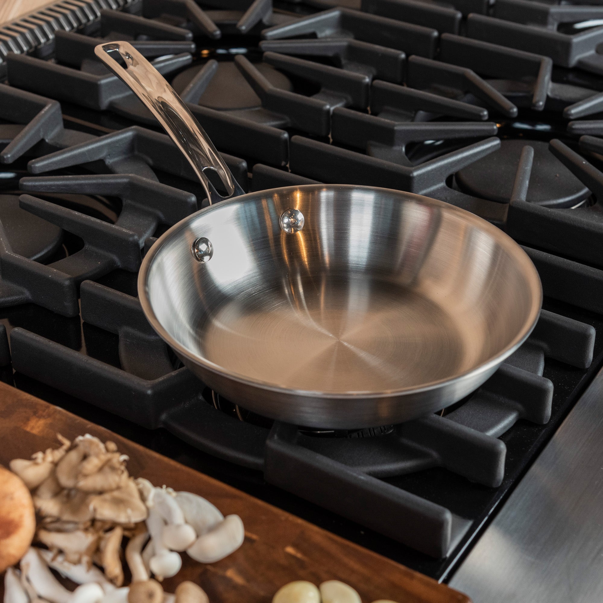 A Viking Culinary Products Professional 5-Ply 8-inch Fry Pan rests on a gas stove with black grates. In front, a wooden board displays mushrooms and garlic, ready to cook. This dishwasher-safe pan streamlines your kitchen routine.