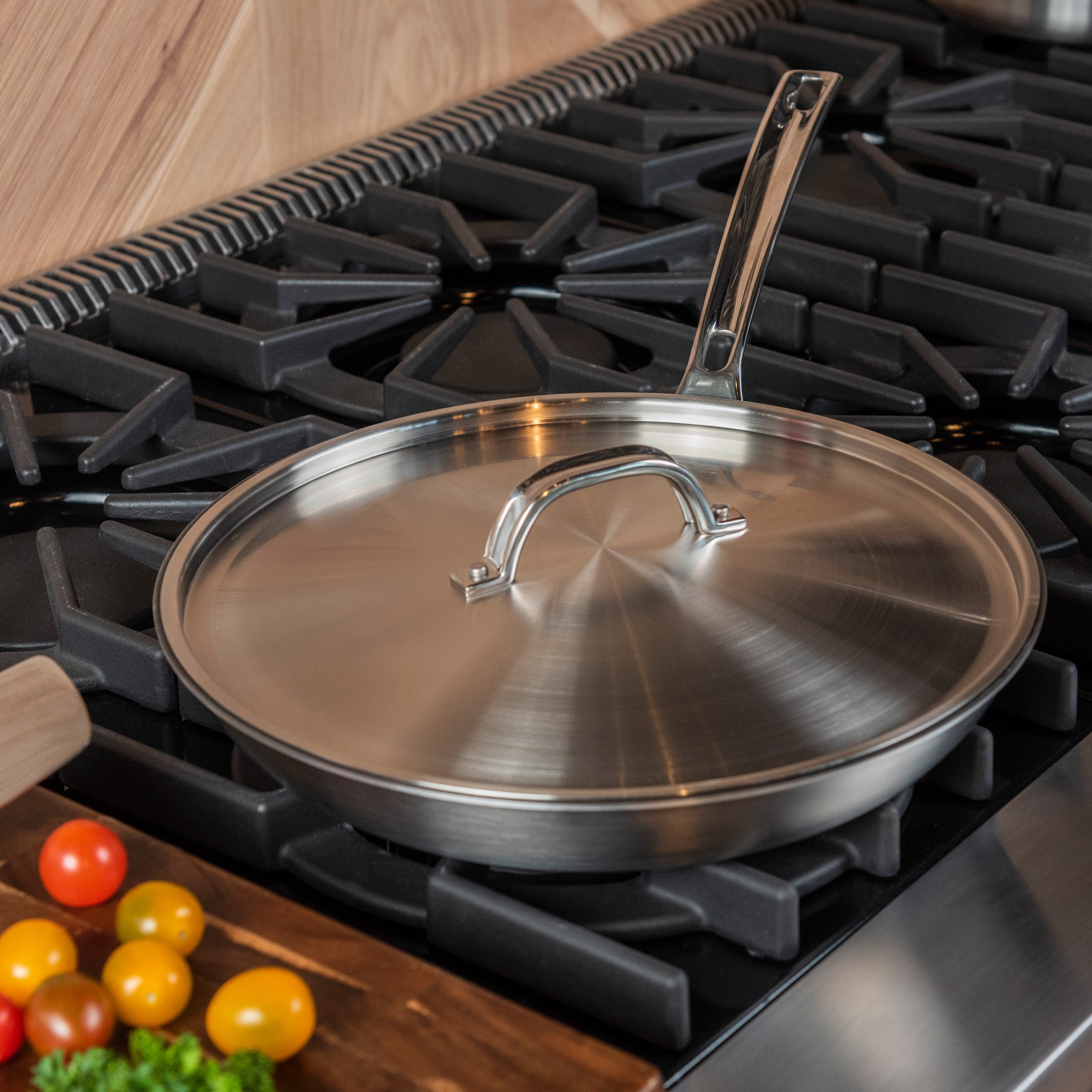 A Viking Culinary Products Professional 5-Ply Stainless Steel Covered Nonstick 12-Inch Fry Pan with Stay-Cool Handles sits on a black gas stove, while fresh tomatoes and parsley are on the wooden countertop nearby.