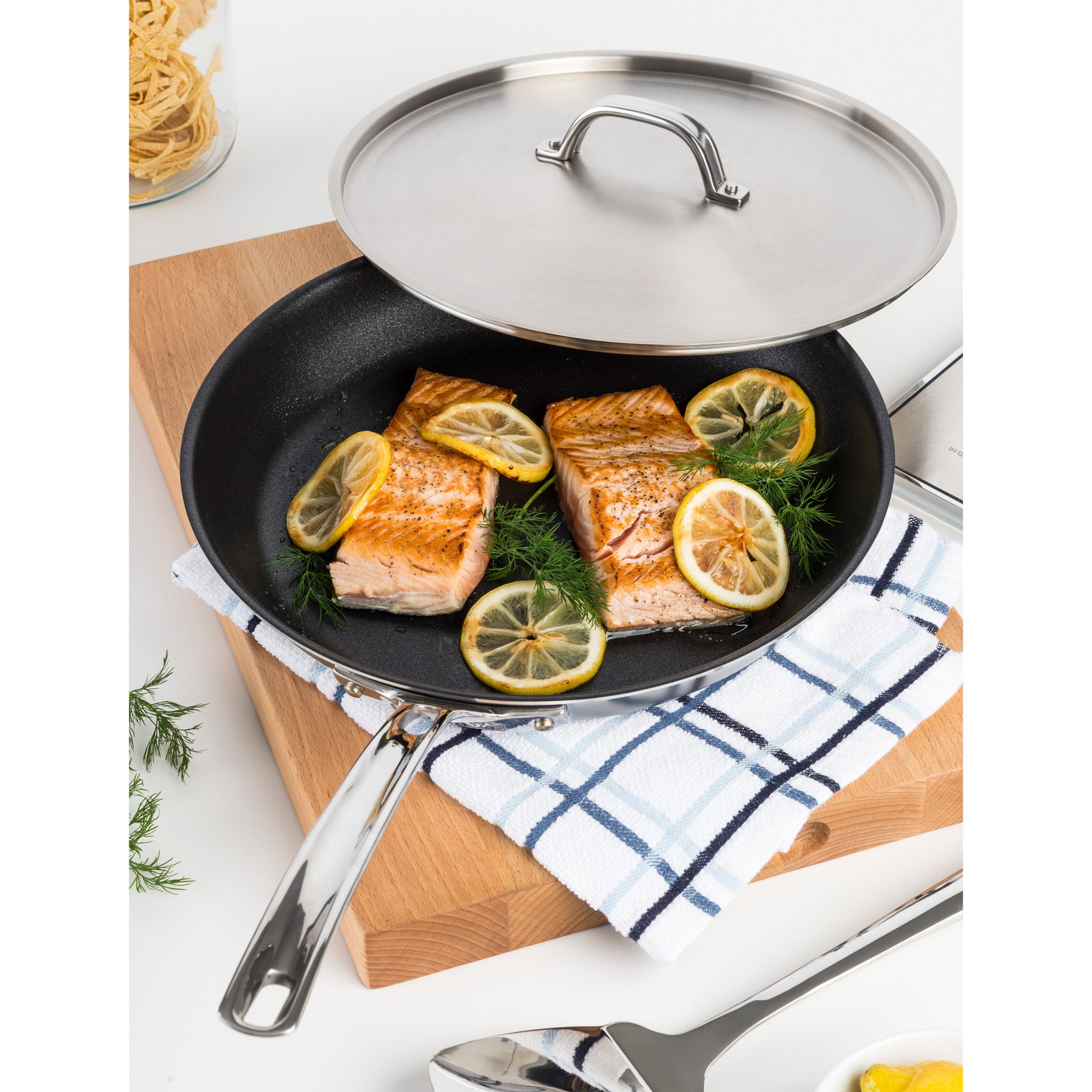 The Viking Professional 5-Ply Stainless Steel Covered Nonstick 12-Inch Fry Pan by Viking Culinary Products holds two salmon fillets with lemon and dill, on a checkered towel and wooden board; the stainless steel lid is partially covering the pan.