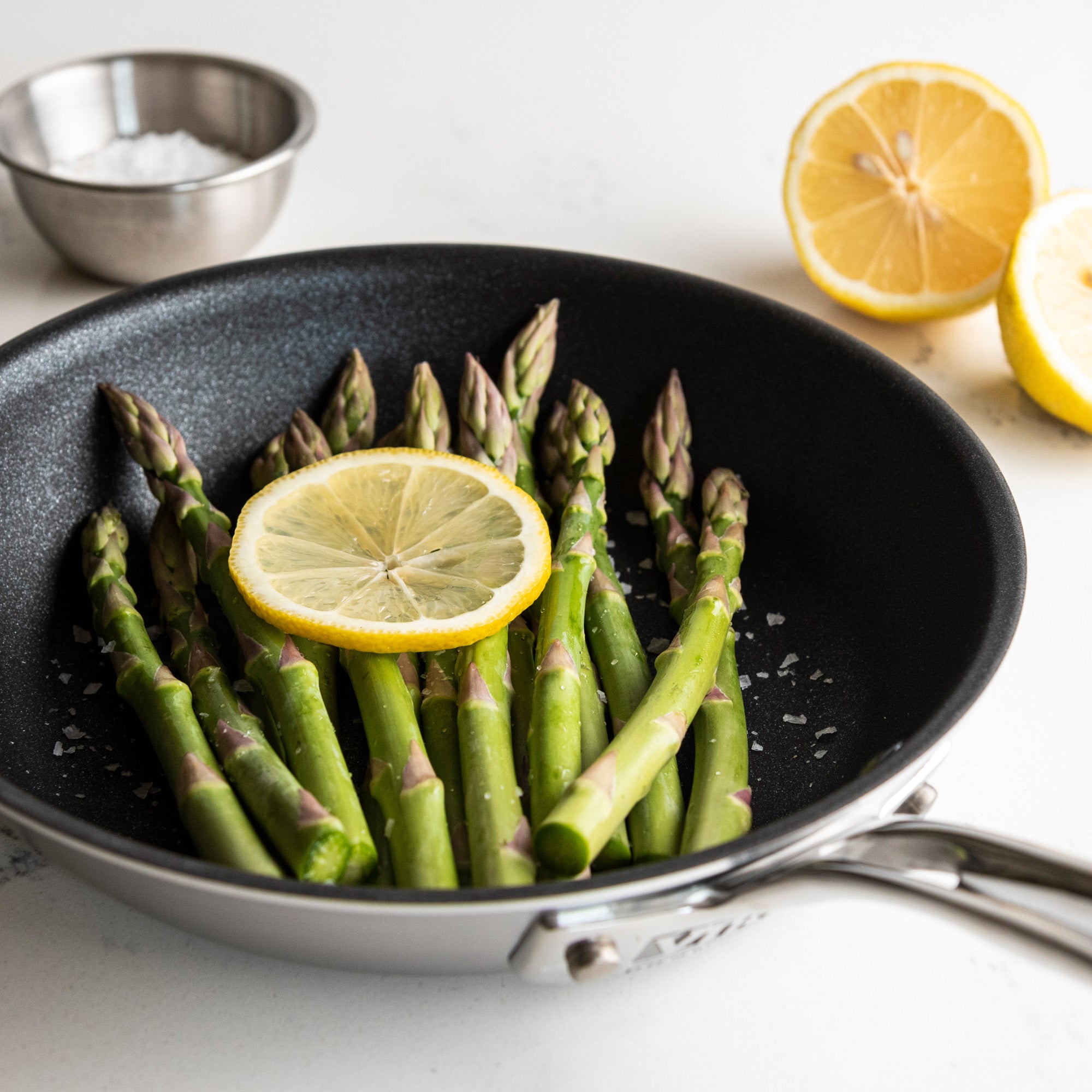 The Viking Professional 5-Ply 8-Inch Nonstick Fry Pan by Viking Culinary Products holds fresh asparagus and a lemon slice with coarse salt. In the background, a metal bowl with salt and half a lemon sit on a white surface.