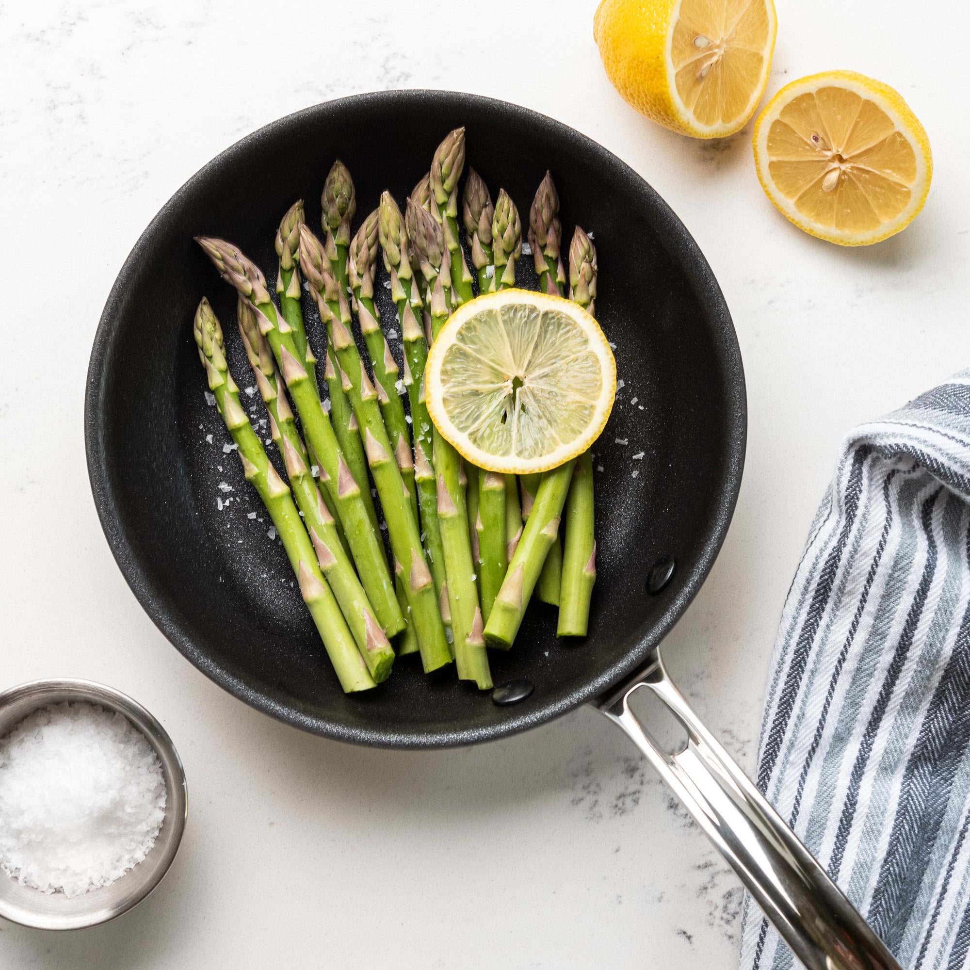 A black Viking Professional 5-Ply 8-Inch Nonstick Fry Pan by Viking Culinary Products holds fresh asparagus and a lemon slice, topped with coarse salt. A halved lemon, bowl of salt, and striped towel sit on the counter near its Stay-Cool Handles.