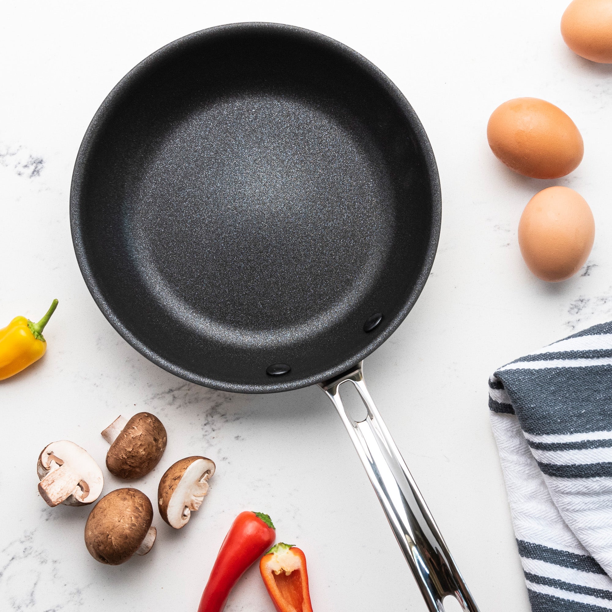 A Viking Culinary Products Professional 5-Ply 8-Inch Nonstick Fry Pan with Stay-Cool Handles rests on a white surface, surrounded by three brown eggs, mushrooms, yellow and red peppers, and a blue-striped kitchen towel.