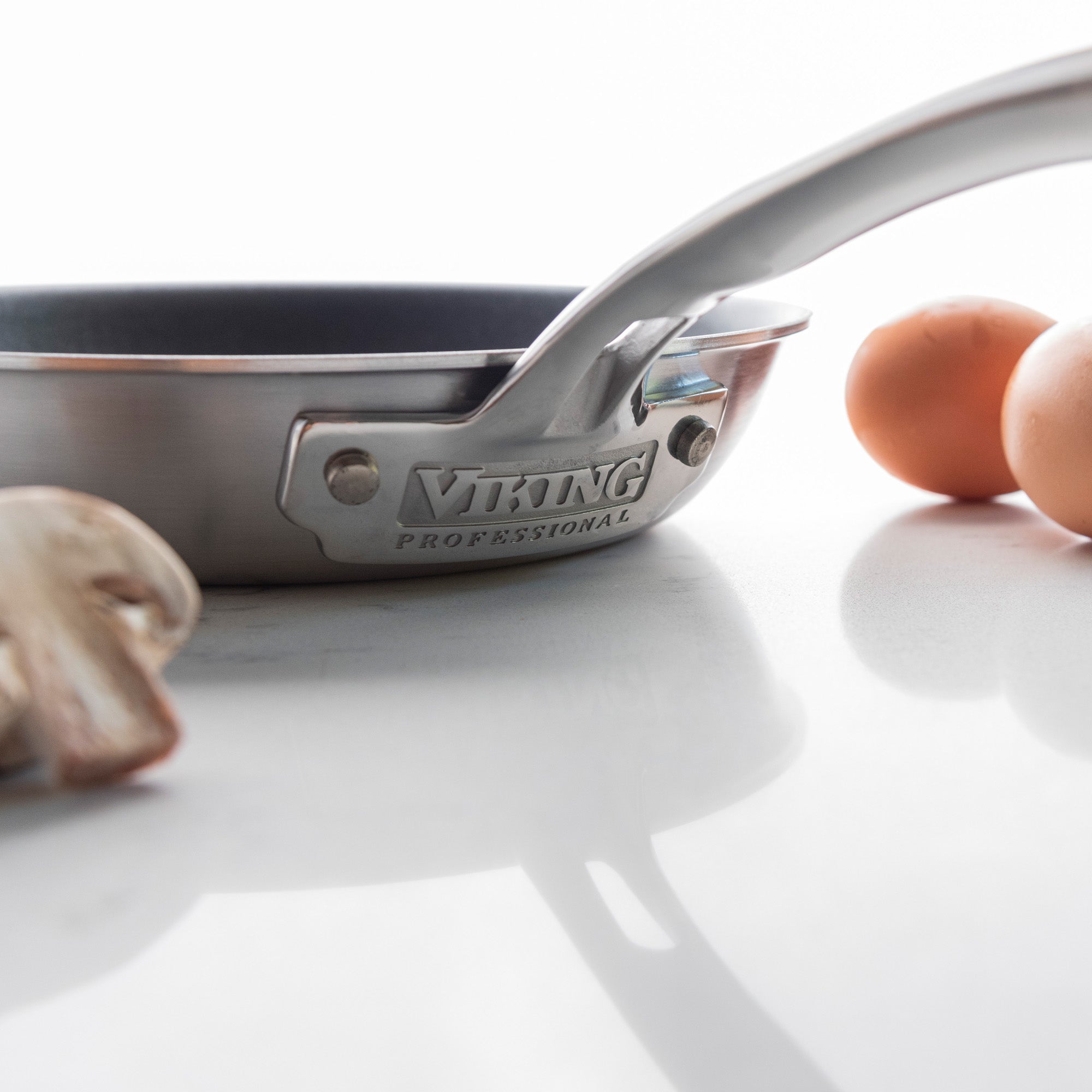 A Viking Professional 5-Ply 8-Inch Nonstick Fry Pan from Viking Culinary Products sits on a white surface, with mushrooms and two brown eggs nearby.