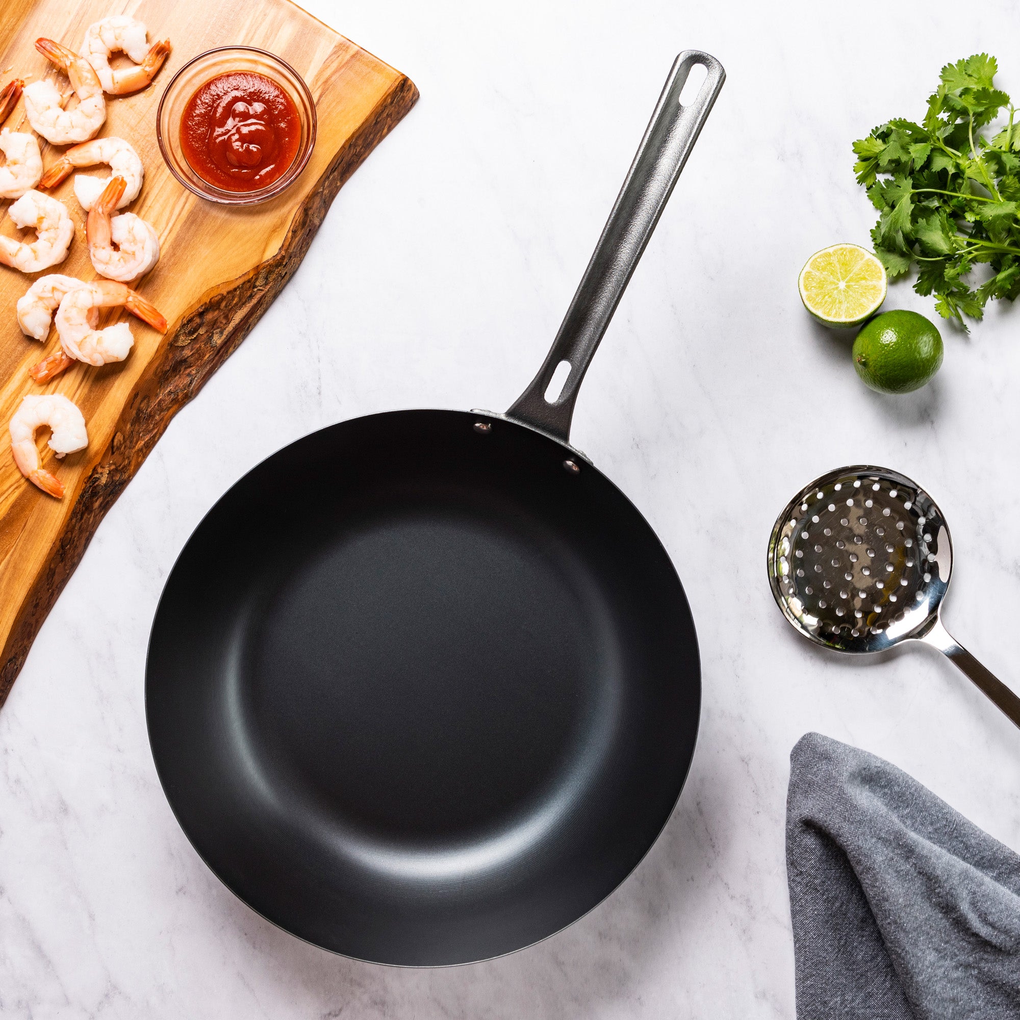 The Viking Culinary Products Blue Carbon Steel 12-Inch Wok sits on a marble surface, surrounded by raw shrimp, a bowl of sauce, halved lime, cilantro, a slotted spoon, and a gray kitchen towel.