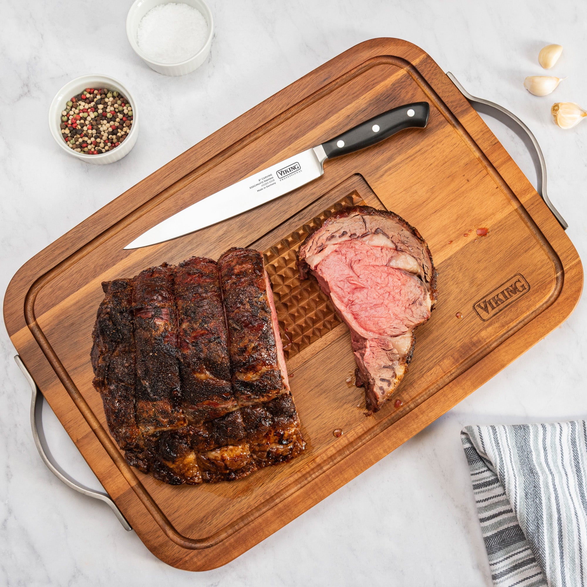 A sliced roast beef rests on the Viking Acacia Carving Board with Juice Well and Metal Handle by Viking Culinary Products. A carving knife, salt, peppercorns, garlic cloves, and a striped towel complete the marble countertop scene.