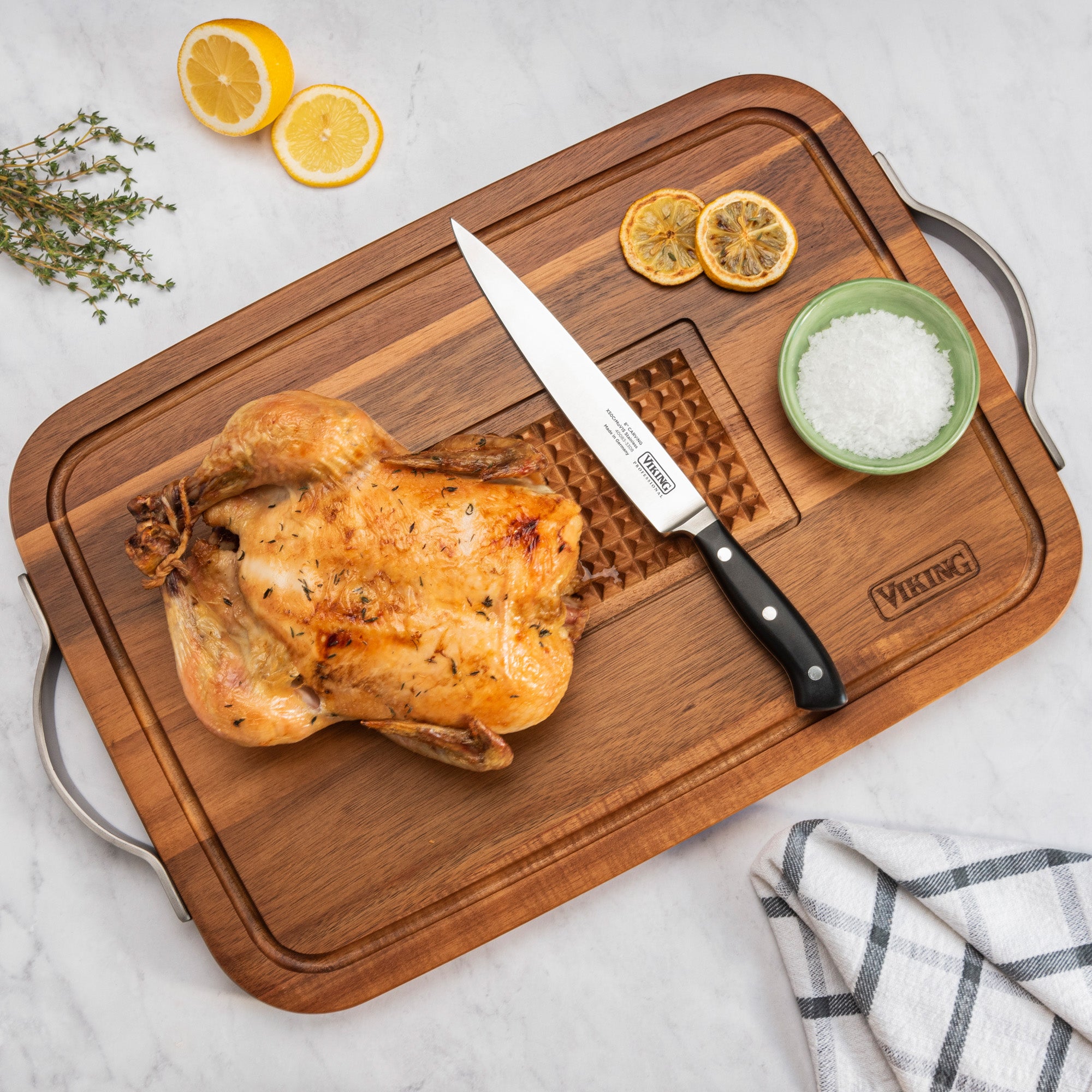 A roasted chicken rests on the Viking Acacia Carving Board with Juice Well and Metal Handle by Viking Culinary Products, accompanied by a chefâ€™s knife, coarse salt dish, lemon slices, fresh thyme, and a kitchen towel on a marble counter.