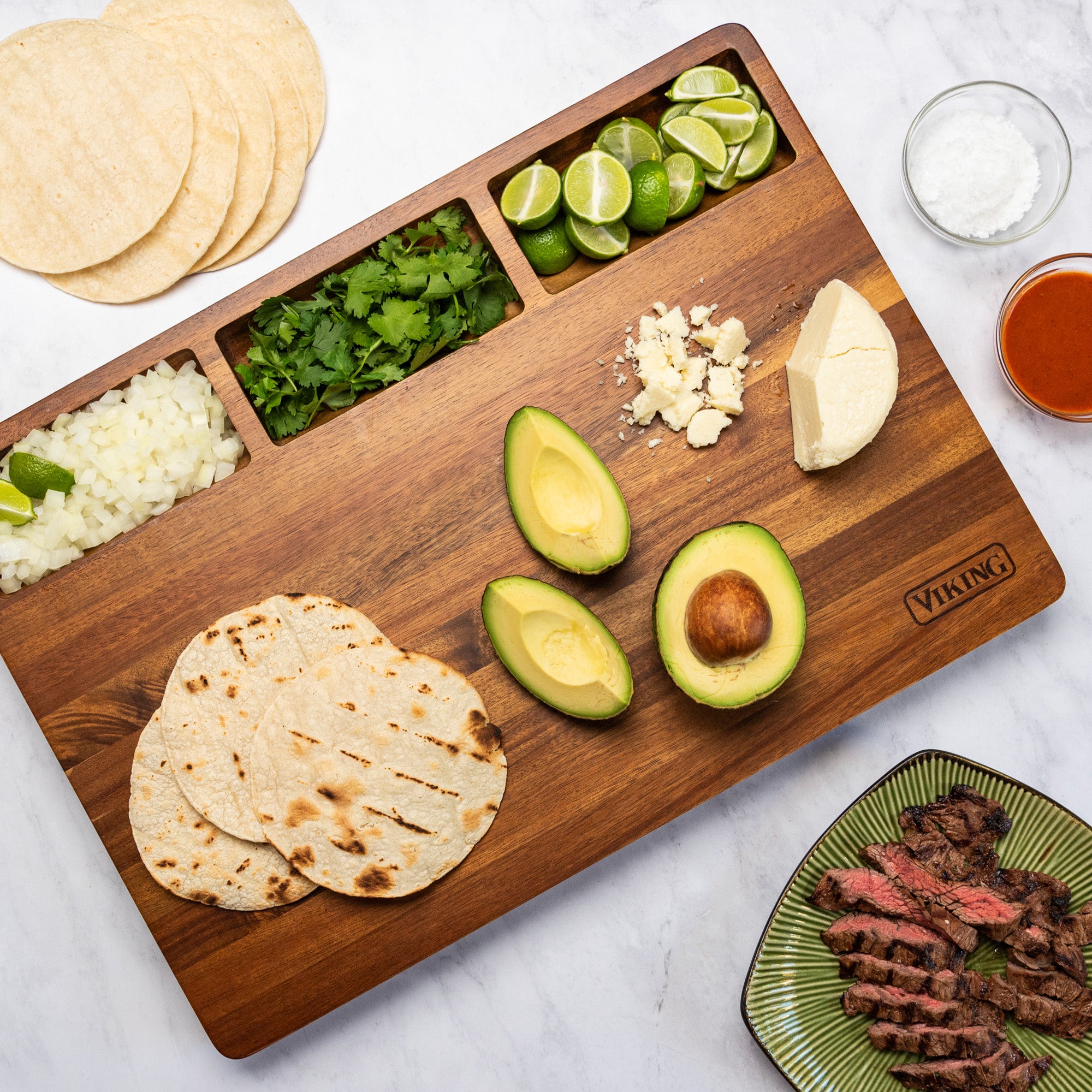A Viking Acacia 20-Inch Reversible Butcher Block Prep and Carving Board by Viking Culinary Products holds tortillas, avocado, cheese, onion, cilantro, lime wedges, and salt. Nearby is a green plate with sliced grilled steak and red sauce.