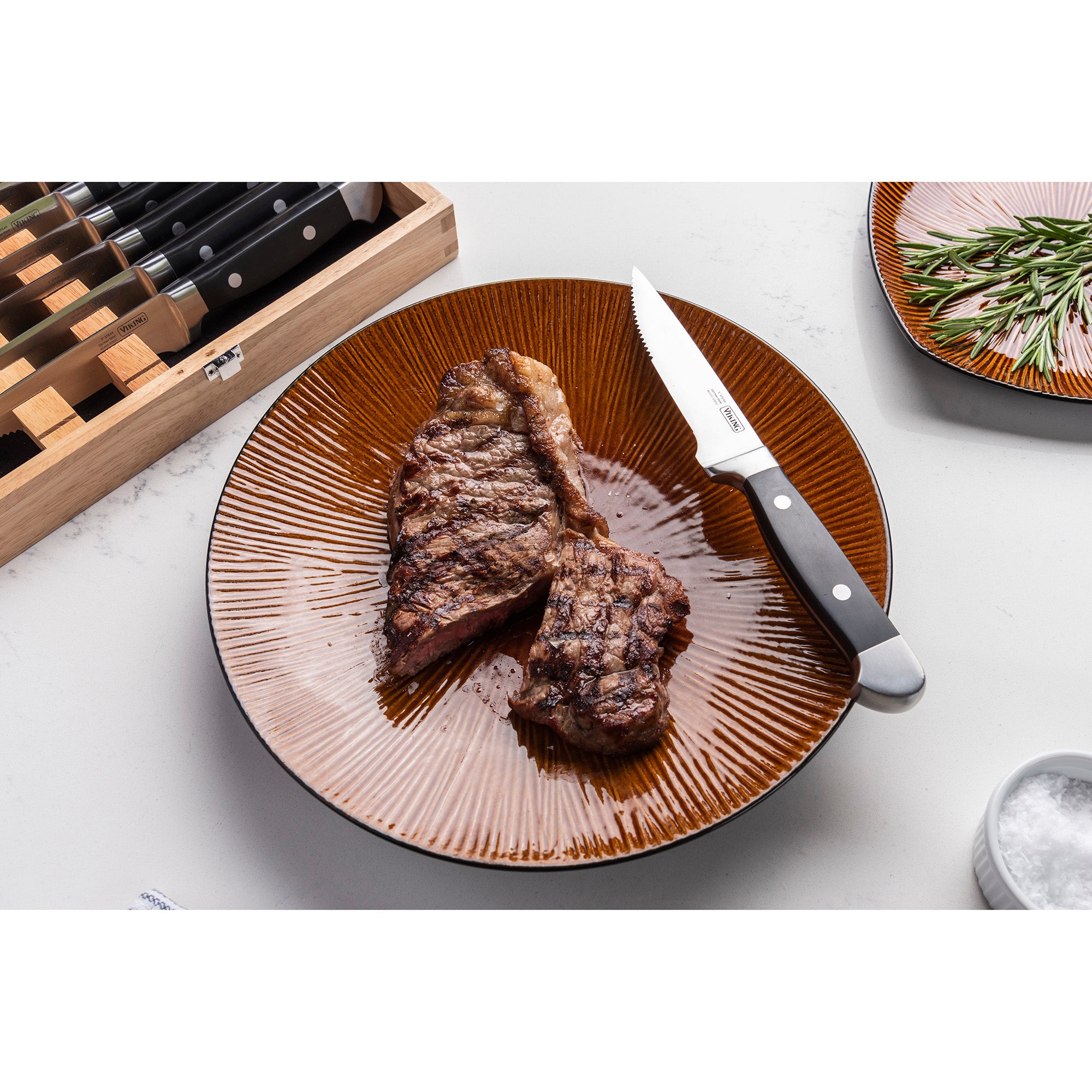 A grilled steak, sliced on a brown plate, is served beside a Viking Steakhouse 8-Piece Steak Knife from Viking Culinary Products. The gift box, rosemary plate, and salt container rest nearby on a white countertop.