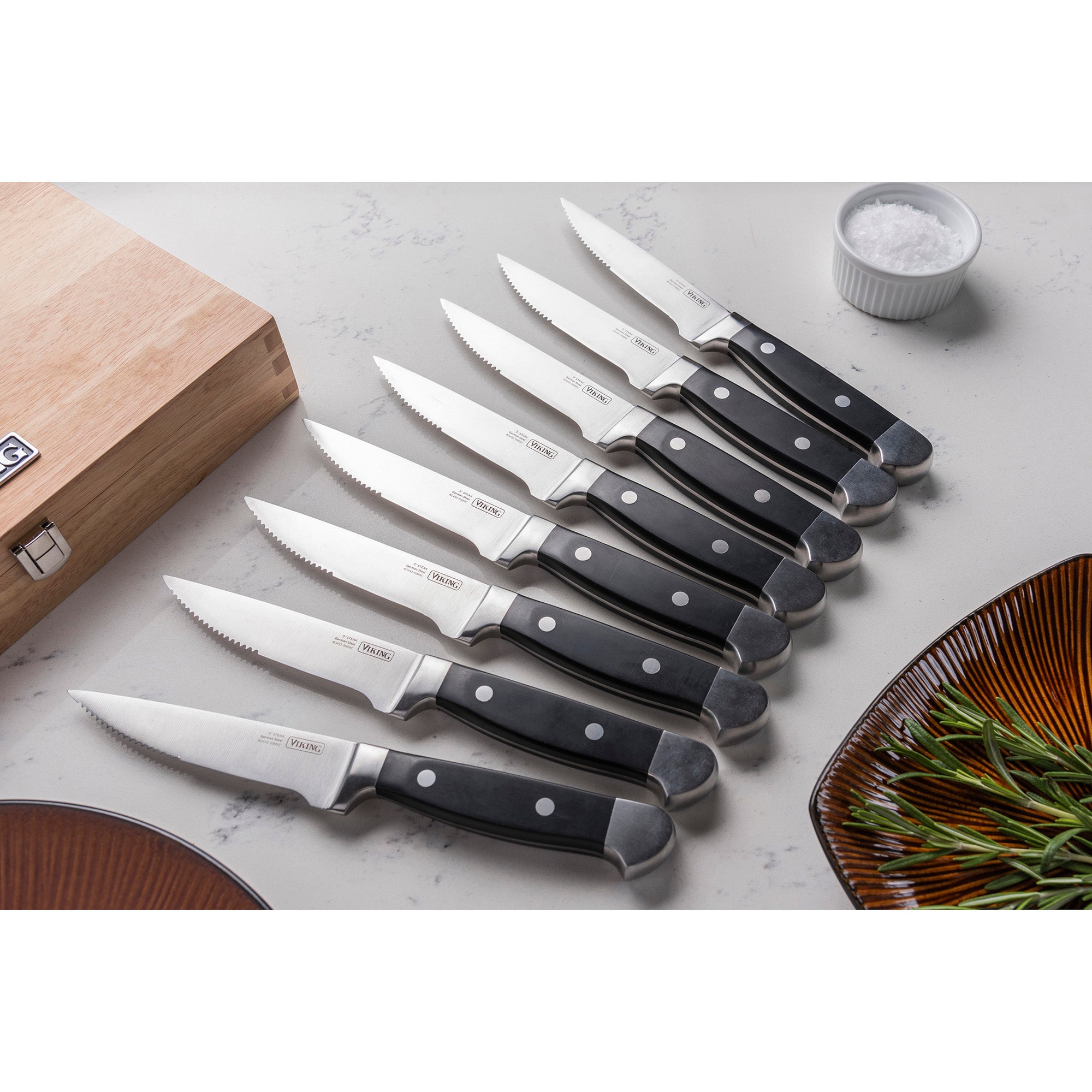 Eight serrated steak knives from the Viking Steakhouse 8-Piece Steak Knife Set by Viking Culinary Products, with black handles, are displayed on a white countertop beside their gift box, a bowl of salt, and fresh rosemary.