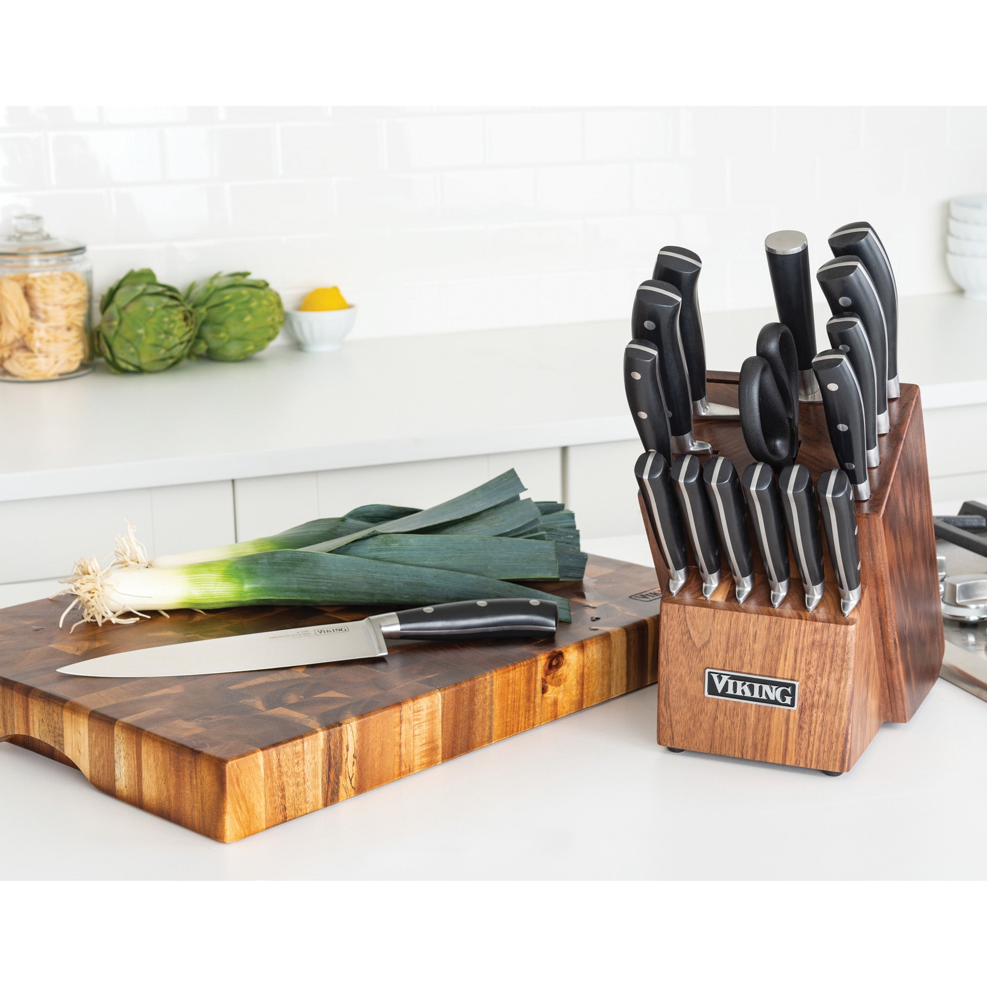 A Viking 17-Piece Cutlery Set with Light Walnut Color Block by Viking Culinary Products sits on the counter next to a chefâ€™s knife, leek, and cutting board, with stainless steel kitchen items and vegetables in the background.