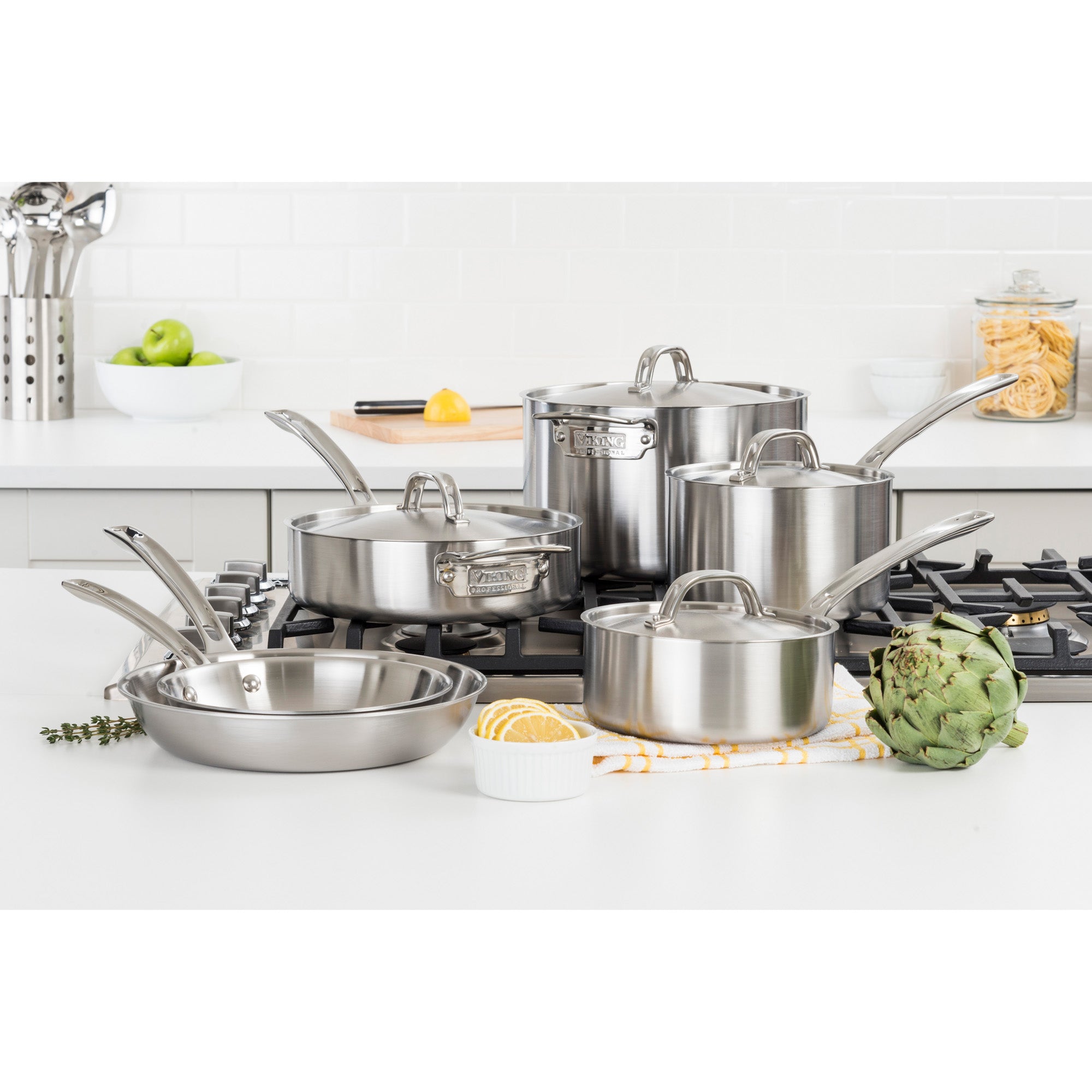 The Viking Professional 5-Ply 10-piece Cookware Set from Viking Culinary Products is shown on a stovetop in a modern kitchen, surrounded by fresh vegetables, utensils, and other kitchen items in the background.