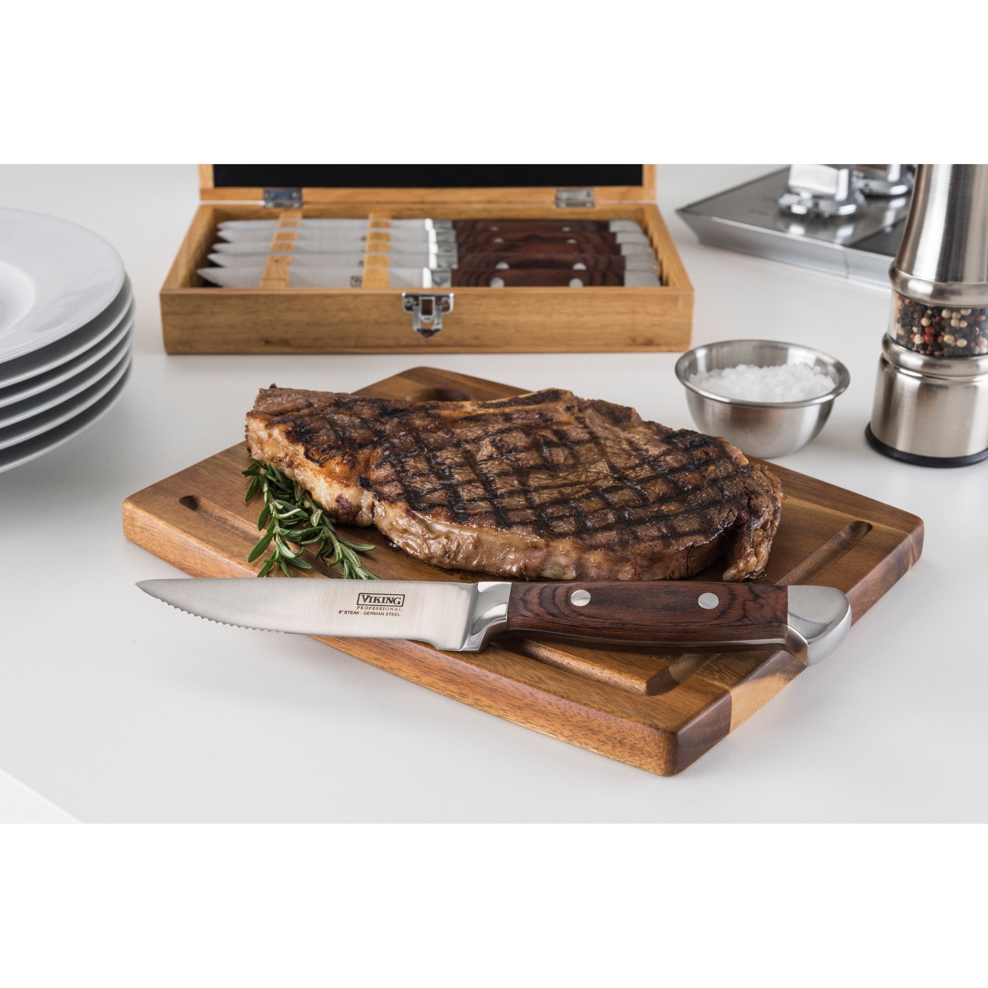 A grilled steak sits on a cutting board next to a Viking Steakhouse Pakka Wood Steak Knife by Viking Culinary Products; in the background are salt, a pepper grinder, stacked plates, and the red 6-piece knife setâ€™s gift box.