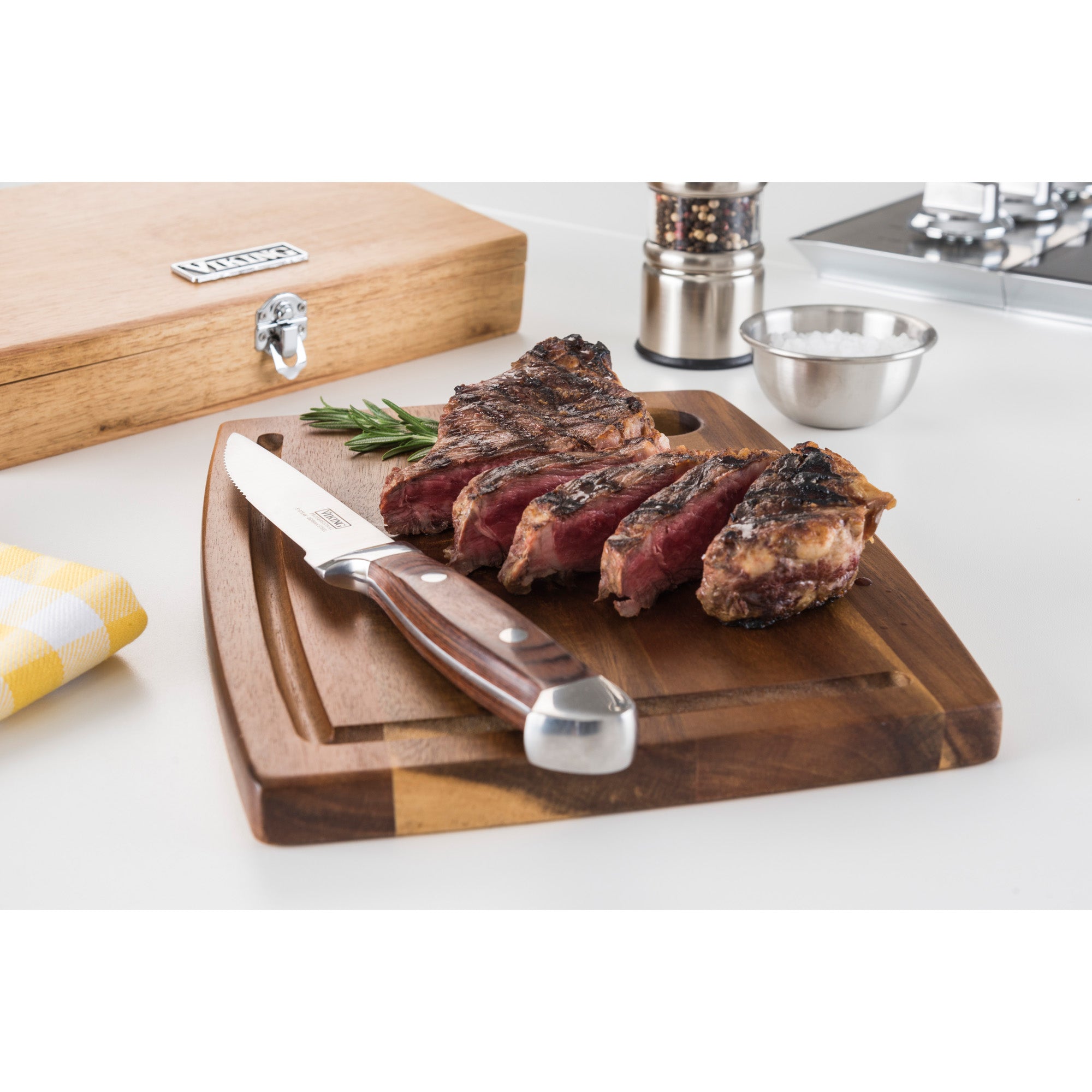 Sliced grilled steak on a wooden board with a Viking Steakhouse Pakka Wood 6-Piece Steak Knife Set by Viking Culinary Products, rosemary, salt bowl, pepper grinder, and the red gift box displayed in a bright kitchen.