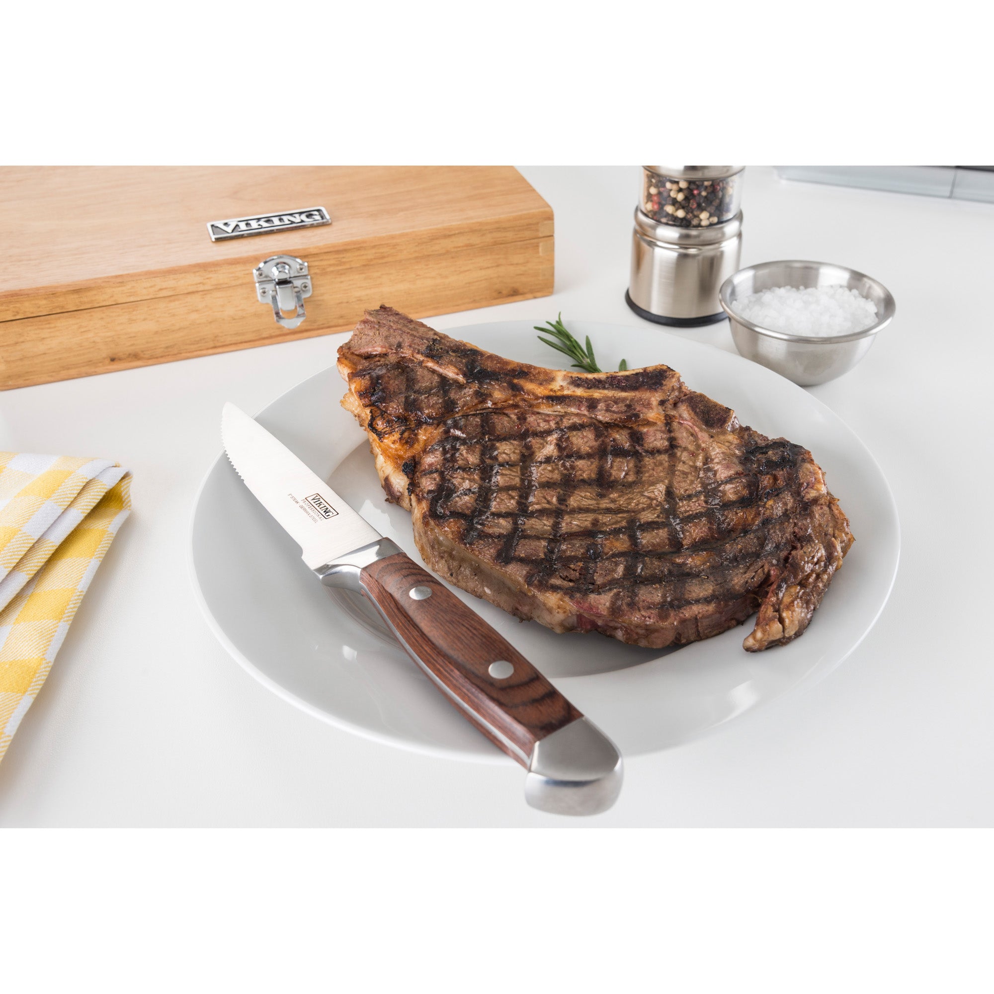 A grilled steak with grill marks sits on a white plate beside a Viking Steakhouse Pakka Wood Steak Knife from Viking Culinary Products. Nearby are a yellow-checked napkin, red gift box, pepper grinder, bowl of salt, and rosemary sprig.