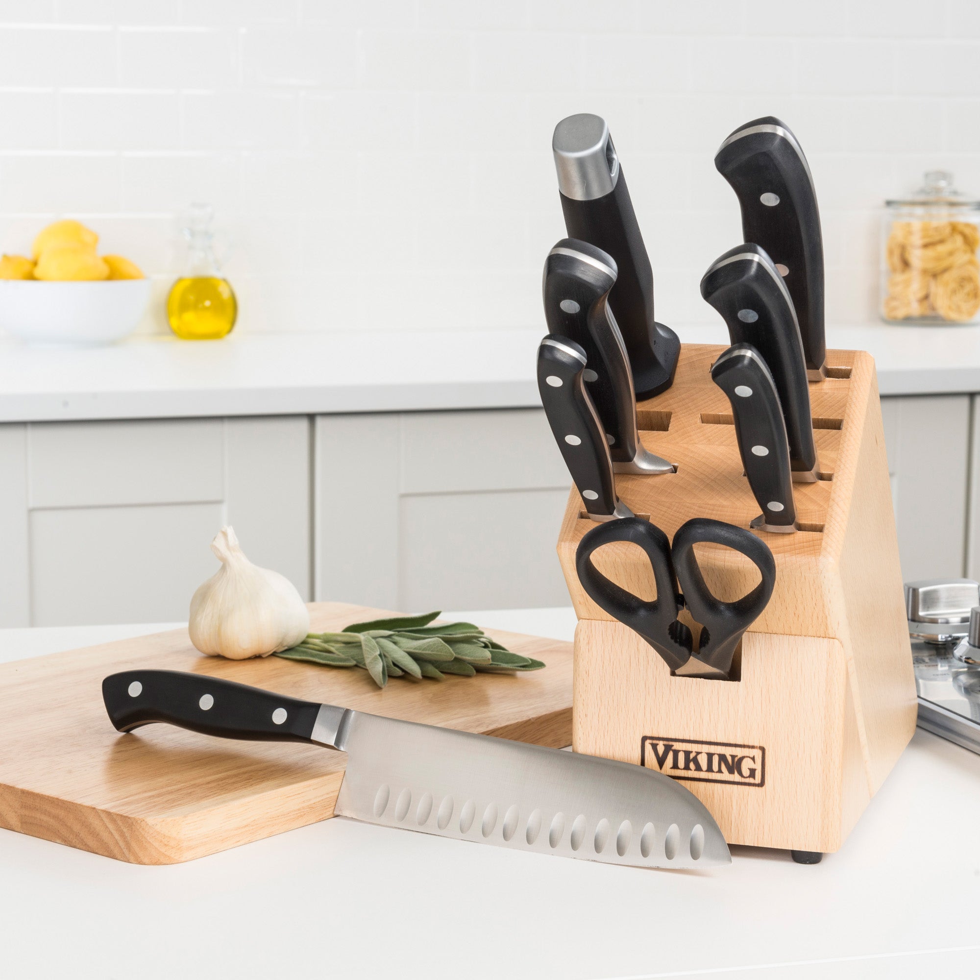 The Viking Professional 9-Slot Beechwood Knife Block by Viking Culinary Products holds several kitchen knives and scissors on a white counter; a chefâ€™s knife rests on a cutting board with garlic, herbs, and lemons nearby. Lifetime Warranty included.